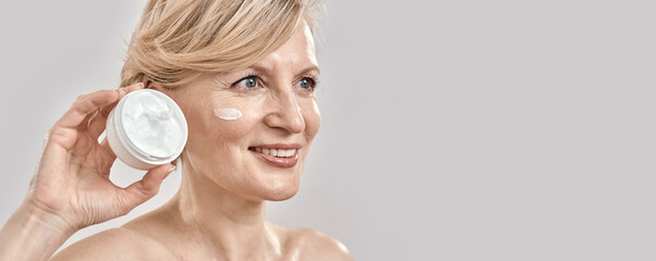 Portrait of beautiful middle aged woman smiling aside, holding cream jar while posing with cream applied on her face isolated over grey background