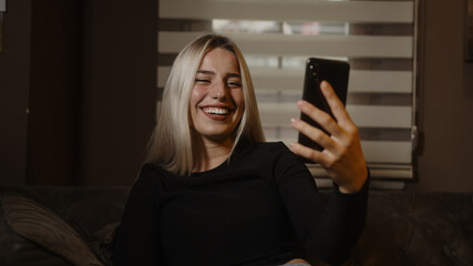 Sweet blonde woman sitting on the sofa while looking at her phone is suddenly surprised, lifestyle, technology, communication and social media concept