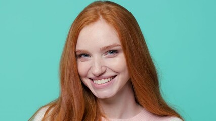 Close up of smiling cheerful ginger redhead young girl in white pink sweater posing isolated on blue turquoise color background in studio. People emotions lifestyle concept. Looking camera blinking - Powered by Adobe