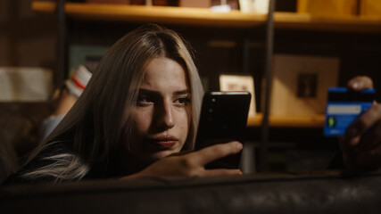 Young blonde beautiful woman sitting on sofa she is shopping with phone and credit card in hand....