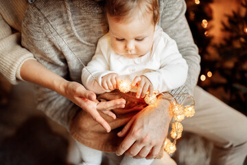 Cute baby girl sit on father caring arms, looking at the light garland, joyful toddler excited about first winter holidays, Christmas and New Year celebration concept