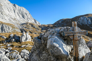 Small, wooden cross on top of a passage in the region of Hochschwab in Austrian Alps. There are massive stony wall in the back. The flora overgrowing the slopes is golden. Spirituality, memory place