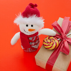 Christmas composition on a red background. A funny snowman holds a gift wrapped in brown paper and tied with a red ribbon. A lollipop is attached to the gift. Christmas and New Year concept