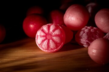 A dark portrait of some pieces of red and pink hard and round candy lying on a wooden table. The main piece of candy has a flower on it, others are just ball shaped.