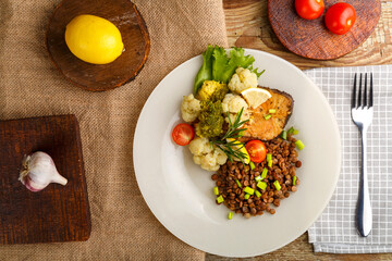Baked fish with broccoli and lentils in a white plate with rosemary and lemon on a checkered napkin next to a fork.