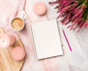 Top view of a notebook with a blank page, a pen, pink candles, a wooden plate, a cup of coffee and heather flowers on pink marble table
