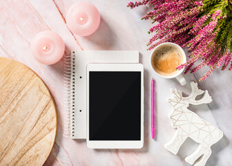 Top view of a empty tablet, a pen, pink candles, a wooden plate, a cup of coffee and heather flowers on pink marble table
