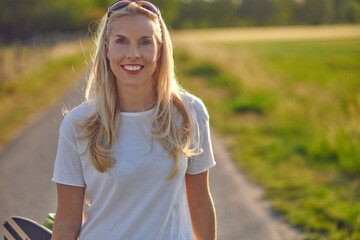 Portrait of a fit beautiful middle-aged woman with an active lifestyle smiling and looking at camera while holding a longboard on a sunny road in the park in summer