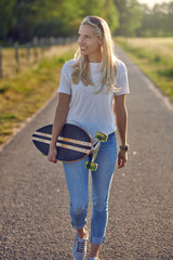 Fit healthy young blond woman carrying a skateboard along a narrow country road looking off the side with a happy smile backlit by the sun
