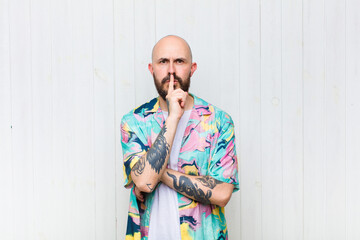 bald man looking serious and cross with finger pressed to lips demanding silence or quiet, keeping...