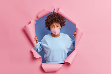 Frustrated unhappy curly haired young woman has sad tired face expression being ill with coronavirus looks displeased at camera feels lonely and stays at home poses against pink torn background