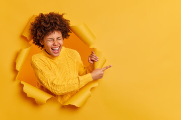 Funny brunette joyful woman with Afro hair wears basic yellow sweater points index finger at blank space and laughs happily stands overjoyed in torn background gestures over yellow studio wall