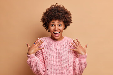 Excited cheerful dark skinned woman raises palms and smiles broadly has surprised face expression wears casual knitted jumper expresses positive emotions poses against brown studio background