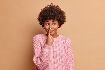Beautiful dark skinned young woman with Afro hair points at nose with index finger has glad expression foolishes around and looks away dressed in knitted sweater isolated over brown background