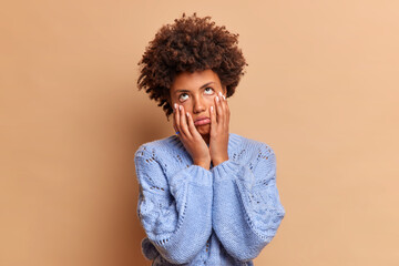 Fototapeta na wymiar Bored frustrated ethnic woman touches face has fatigue expression being sick and tired of weekdays needs rest dressed in blue sweater isolated over brown background. Negative feelings concept