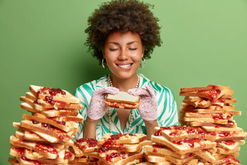 Pleased young female model with Afro hair closes eyes and bites lips holds appetizing bread toast...