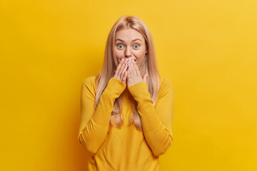 Attractive surprised Caucasian woman with blonde hair covers mouth and stands speechless indoor...