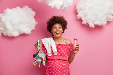Positive African American pregnant woman with curly hair looks above smiling holds feeding bottle mobile toy and carries bodysuit anticipates for baby birth isolated over rosy studio background