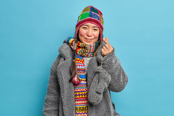 Adorable eastern Asian girl dressed in winter clothing makes korean like sign shows mini heart expresses love going to have walk poses against blue studio background. Valentine concept. Romantic mood