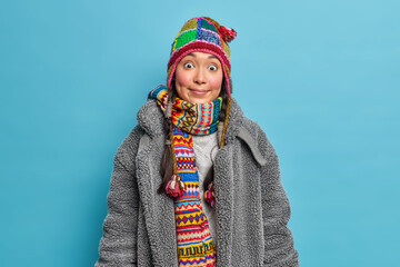 Horizontal shot of surprised young Asian woman with rosy cheeks wears knitted hat scarf around neck and warm fur coat dresses for cold weather isolated over blue background. Winter time concept