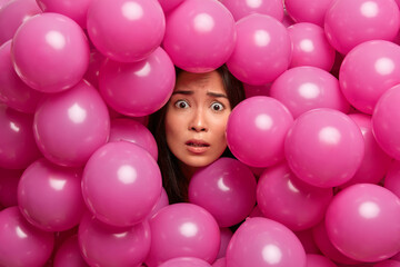 Fototapeta na wymiar Puzzled nervous brunette woman looks embarrassed at camera has worried expression before important event in her life surrounded by small pink inflated balloons. Festive day or holiday concept