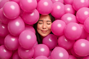 Fototapeta na wymiar Photo of pleased young Asian woman stands with closed eyes smiles gently looks from out inflated pink balloons happy to receive congratulations from close people on her birthday. Festive event