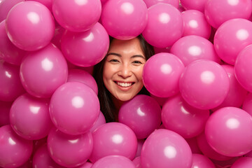 Fototapeta na wymiar Smiling sincere Asian woman sticks out head through pink inflated balloons expresses positive emotions spends free time on party celebrates anniversary or engagement. Holiday decoration concept