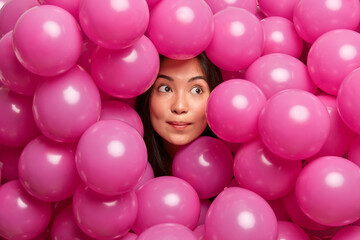 Fototapeta na wymiar Bright shot of thoughtful young Asian woman concentrated aside going to celebrate festive event surrounded by inflated helium pink balloons has birthday party thinks how to entertain guests.