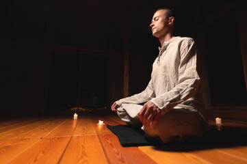 Fototapeta premium Attractive Caucasian young man meditates with closed eyes while sitting in lotus position in a dark room by candlelight.