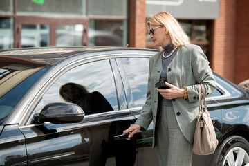 Fototapeta na wymiar Elegant mature businesswoman in sunglasses and grey suit standing by black car
