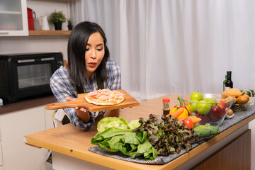 Young attractive Asian woman standing behind wooden kitchen table with different type of vegetables and fruits, holding a wooden tray with pizza and looking at pizza with yummy facial expression.