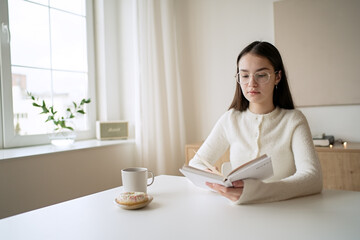 Pretty teenage girl reading book and drinking coffee at home   