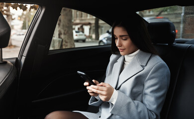 Businesswoman travelling by car on backseat, reading text message on smartphone while driving on meeting