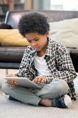 Serious elementary schoolboy of African ethnicity sitting by black leather couch