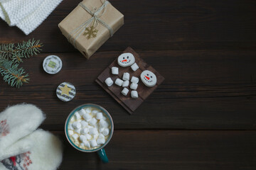 Obraz na płótnie Canvas Christmas New Year card. Cup of hot drink with marshmallows, warm white mittens, New Year's gift, candles on a dark wooden background. Festive concept. Top view. Copy space.