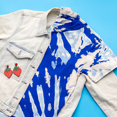Tie-dye t-shirt, denim jacket and strawberry earrings on blue background. Summer fashion style concept