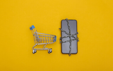 Smartphone wrapped in a steel chain an mini shoping trolley on yellow background. Top view