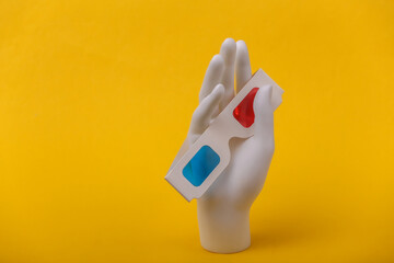 White mannequin hand holding retro anaglyph 3d glasses on yellow background.