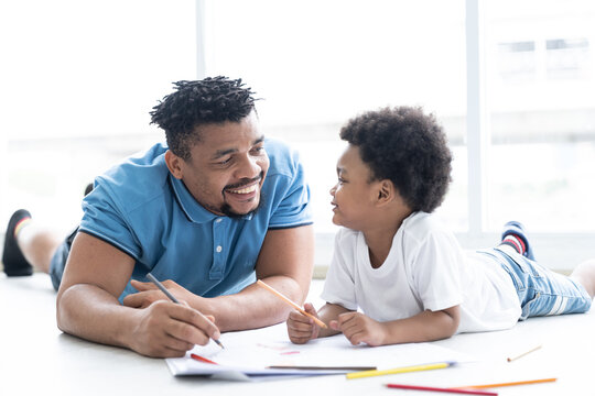 African American smiling father leaning cute curly little son drawing. Happy family lying on floor painting picture together at home. family, kid and education concept