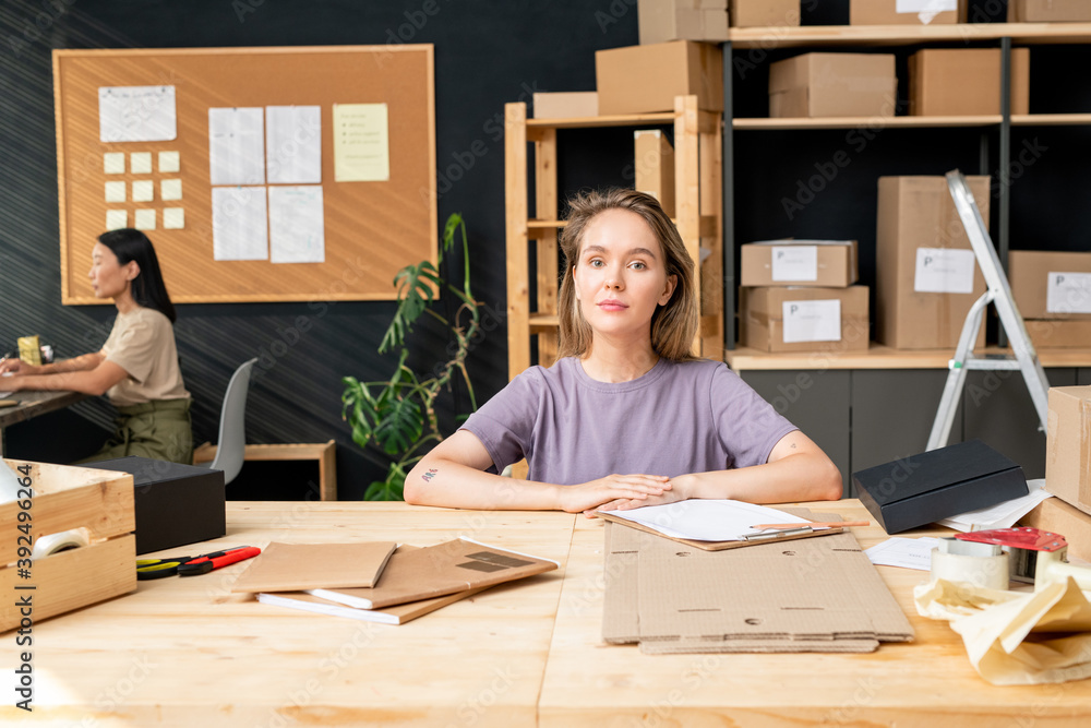 Poster young pretty worker of online shop office looking at you while checking data