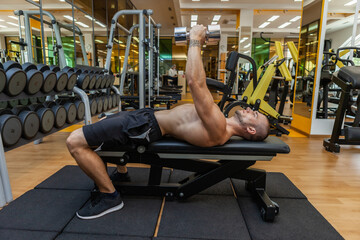 Young muscular fitness man trains pectoral muscles while lying on a bench with dumbbells in his...