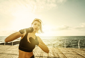 Young woman kickboxer with wrapped hands in bandages trains punch on the beach at sunrise. Morning...