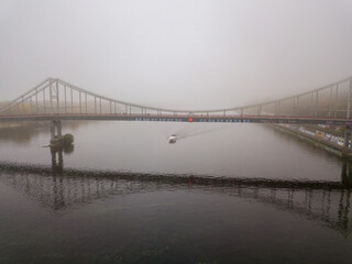 Aerial drone view. A motor boat sails under a footbridge on the Dnieper on a foggy morning.