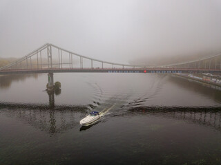 Aerial drone view. A motor boat sails under a footbridge on the Dnieper on a foggy morning.