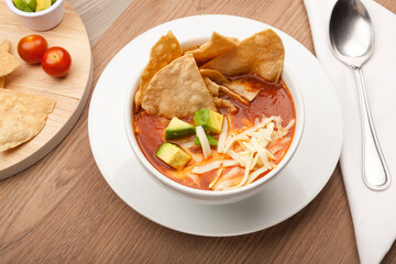 Mexican soup with tortilla chips, avocado and cheese in a white bowl on a rustic wooden table