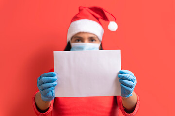 covid19 christmas message. young serious woman holding white blank piece of paper with copy space, santa claus outfit. wearing face mask and plastic gloves. serious expression. red background