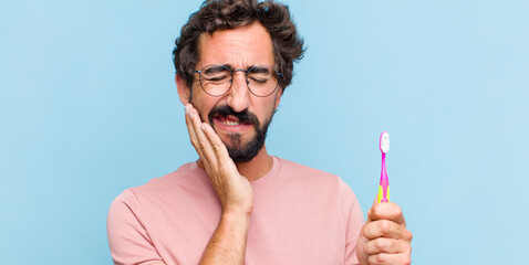 young bearded man holding cheek and suffering painful toothache, feeling ill, miserable and...