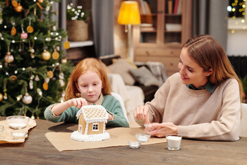 Cute little girl and her mother sprinkling roof of gingerbread house by table