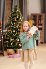 Cute and curious little girl holding giftbox with Christmas present by her ear