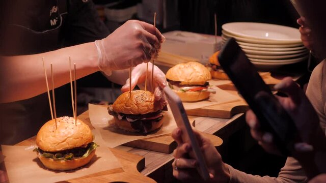 Cooks prepare burgers and cut them on the bar. Chef conducts workshop on cooking burgers. People film the process on their phones